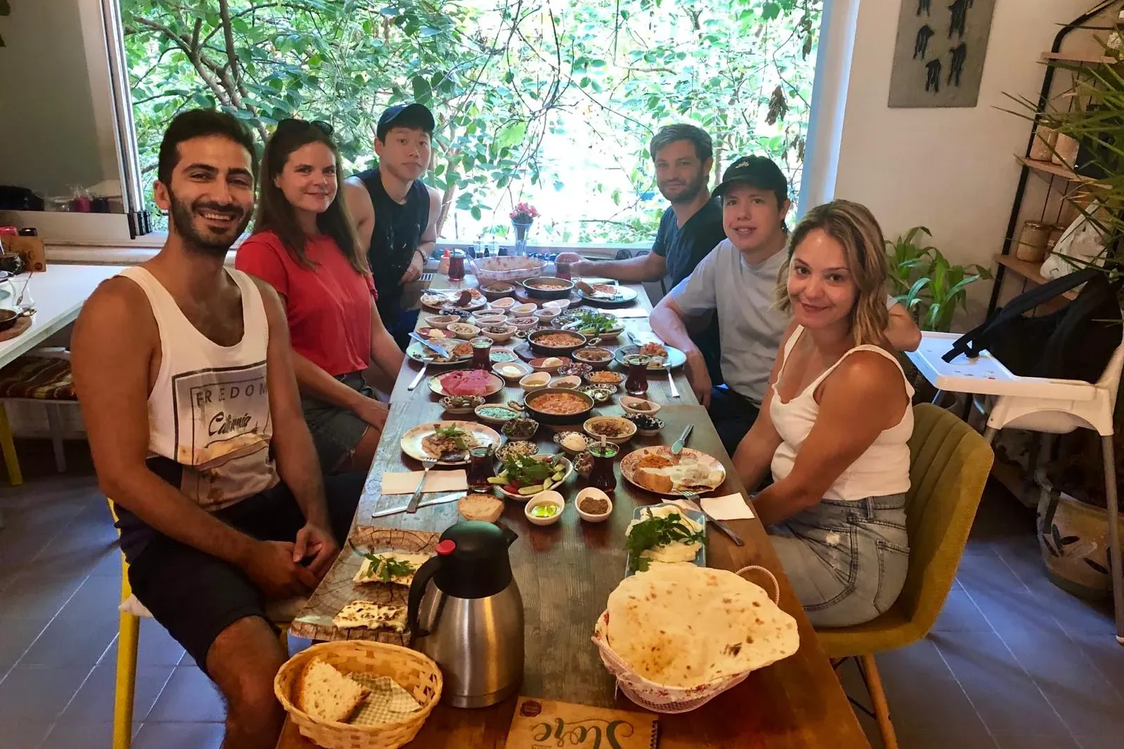 Seis personas sentadas en una mesa larga con varios platos pequeños, pan plano y té, con follaje verde visible al exterior.