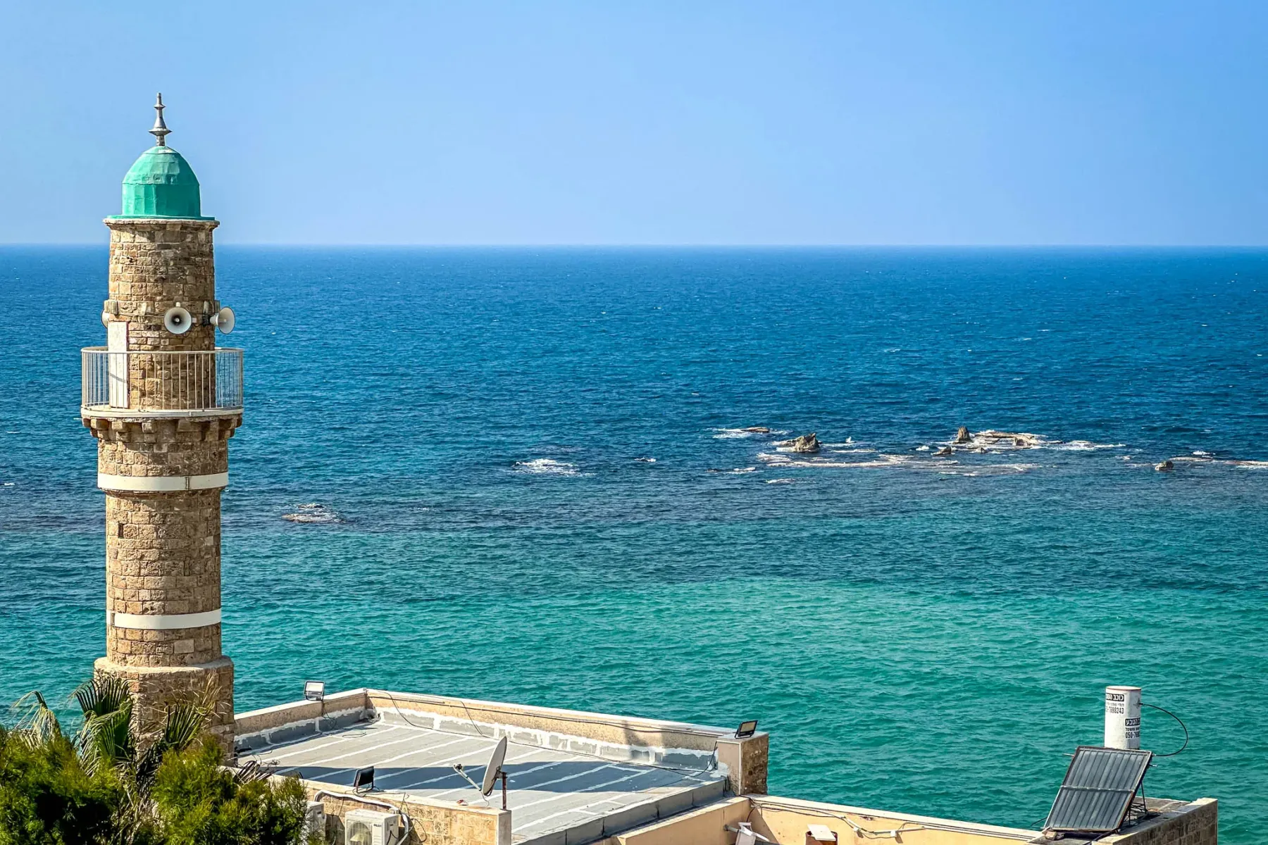 Minarete de piedra con cúpula verde y balcón, dominando el mar Mediterráneo turquesa, con tejados y follaje verde.