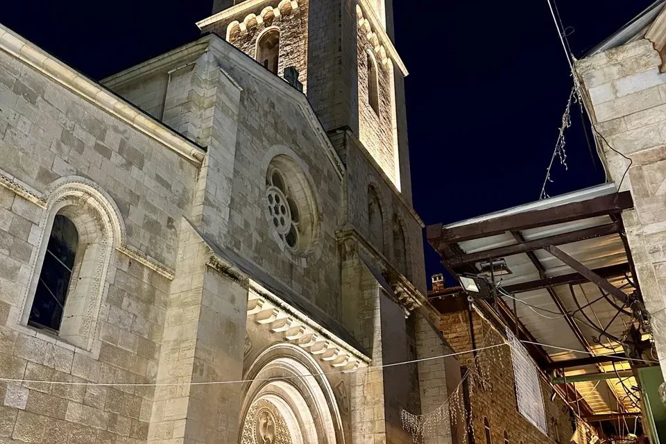 Iglesia de piedra iluminada con alto campanario, entrada arqueada y rosetón de noche en Jerusalén. Edificios adyacentes de...