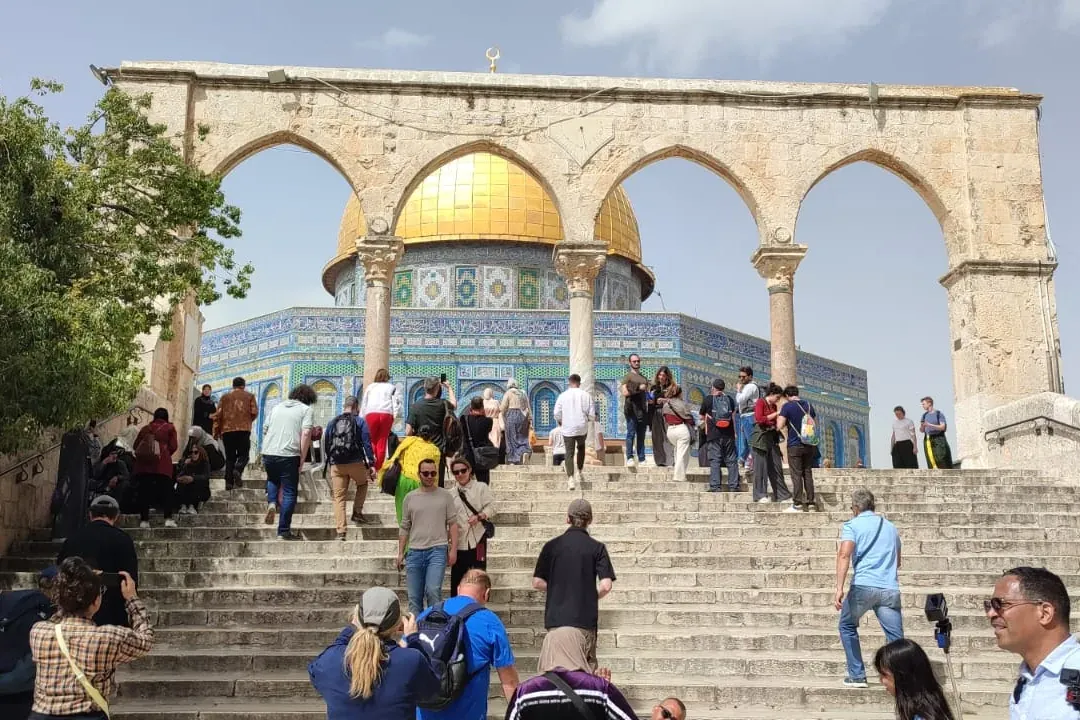 Turistas suben escaleras de piedra hacia la Cúpula de la Roca con cúpula dorada y azulejos azules, bajo arcos de piedra.