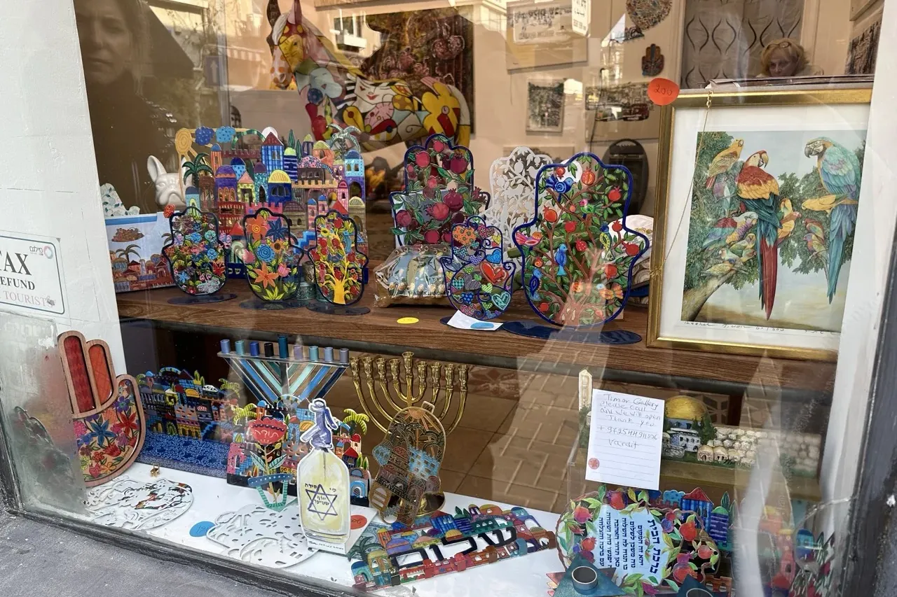 Shop window displaying colorful hamsa hand decorations, menorahs, a painting of parrots, and other traditional souvenirs. ...