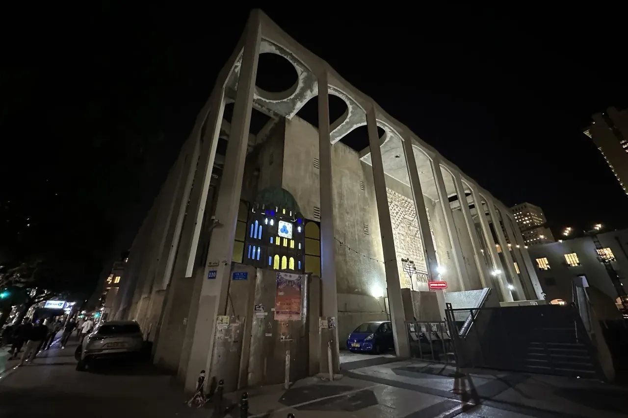 The Great Synagogue of Tel Aviv at night, with its modernist concrete architecture, a street, parked cars, and illuminated...