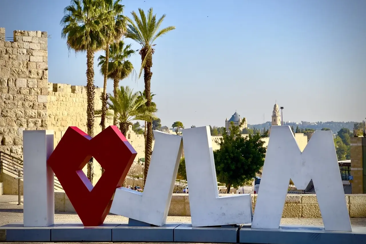 Letras grandes blancas 'I' y 'JLM' con un símbolo de corazón rojo en un espacio público. Detrás hay un muro de piedra, pal...