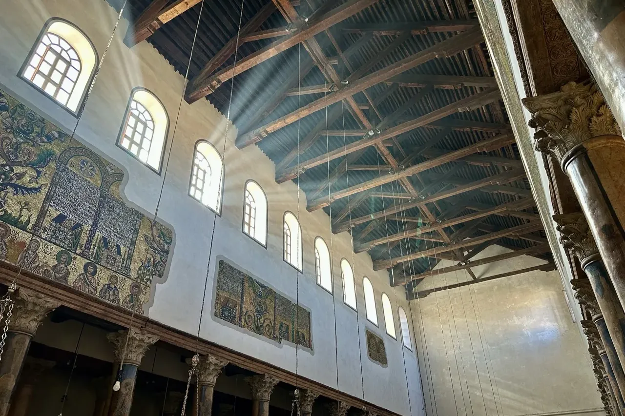 Interior de iglesia con mosaicos dorados antiguos, ventanas arqueadas y techo de madera oscuro iluminado por rayos de sol.