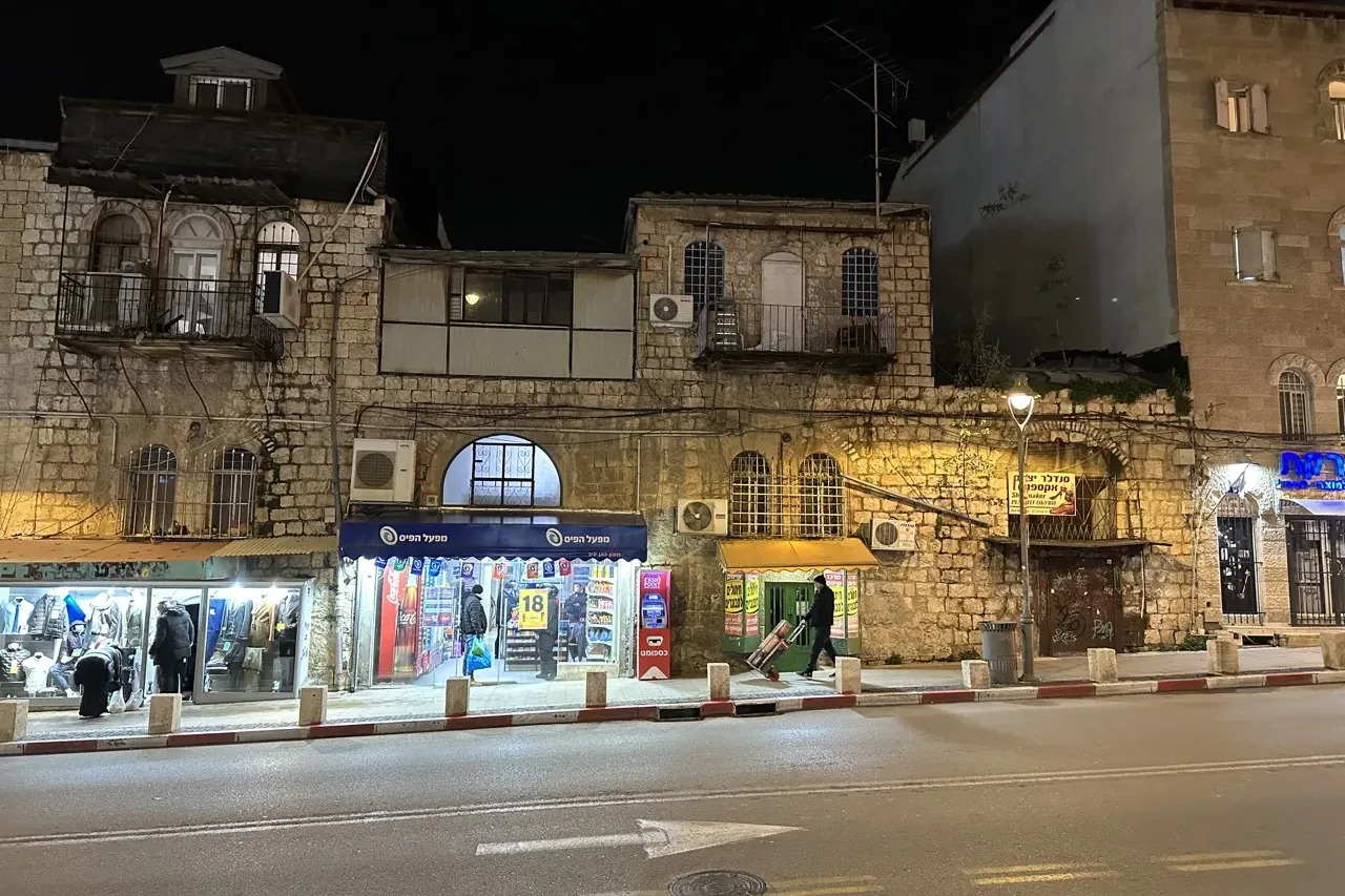 Vista nocturna de una calle en Jerusalén con edificios históricos de piedra, tiendas iluminadas en la planta baja y una pe...