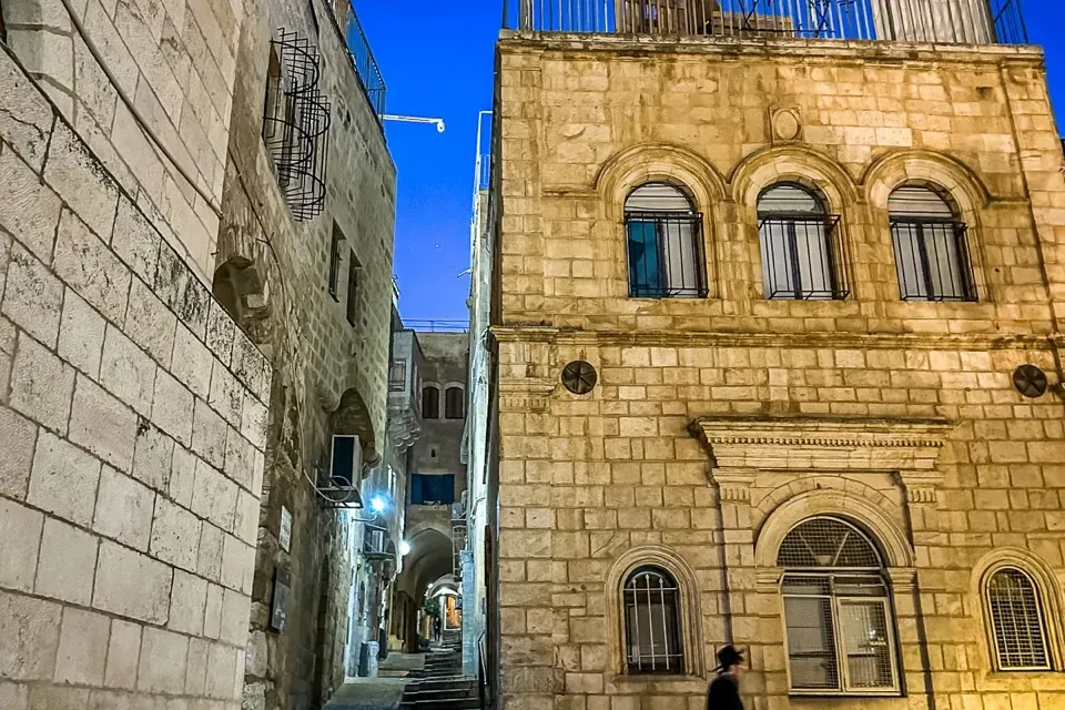 Edificios de piedra flanquean un callejón estrecho y escalonado en Jerusalén por la noche. Una persona baja escaleras a la...