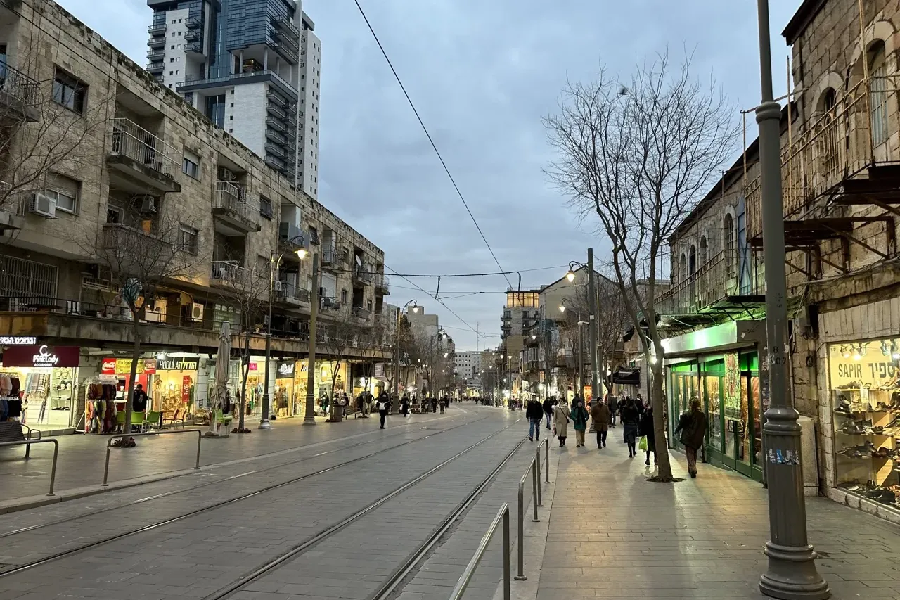 Una calle urbana en Jerusalén al anochecer, con vías de tranvía, edificios de piedra con tiendas, peatones en las aceras y...
