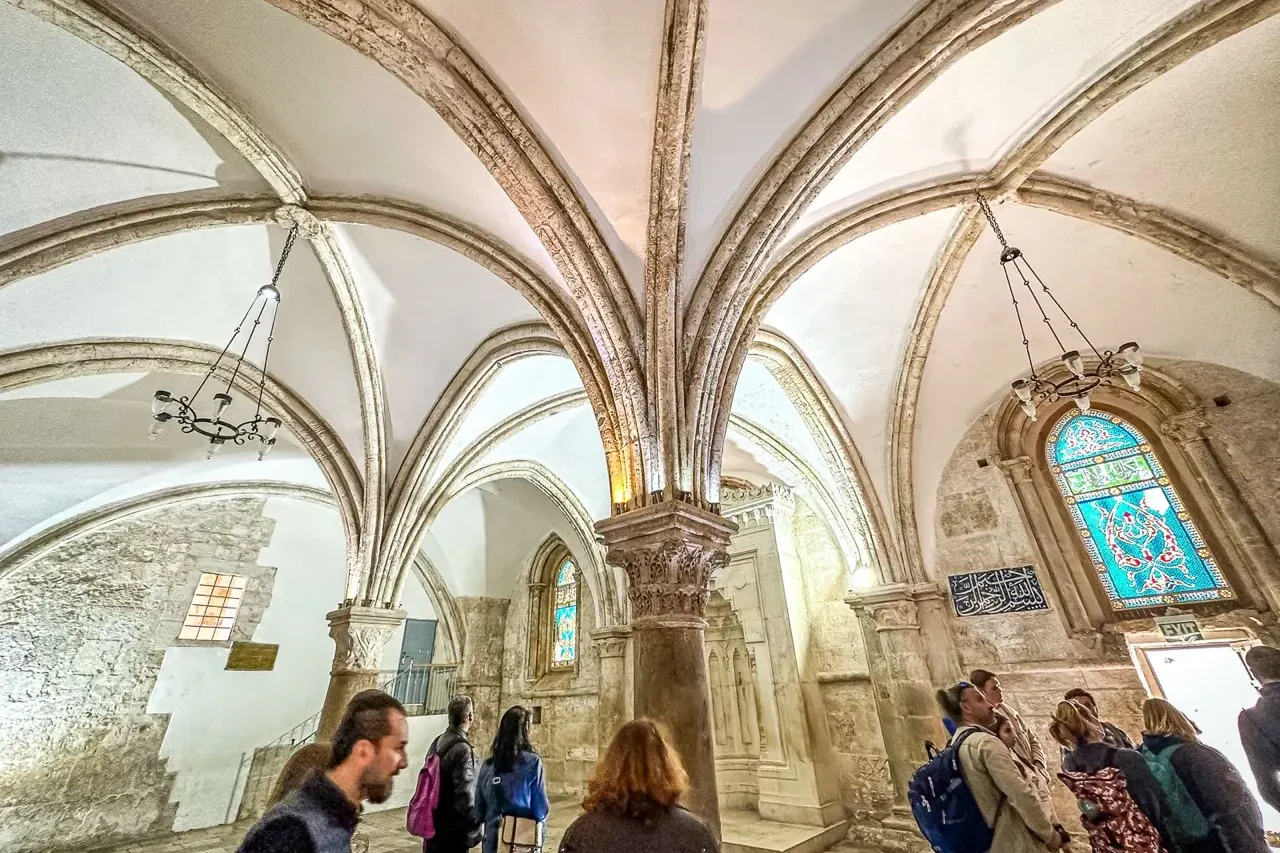 Grupo de turistas en sala abovedada histórica con muros de piedra, techo de nervaduras, vidriera azul y escritura árabe.