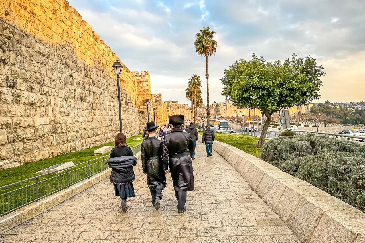 Personas caminan por un sendero adoquinado junto a las murallas de la Ciudad Vieja de Jerusalén, con palmeras y cielo nubl...