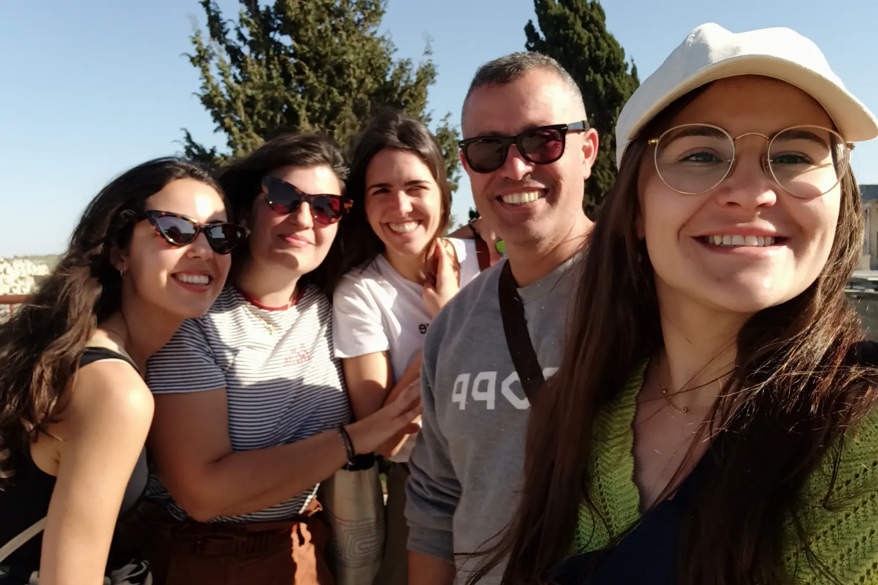 Cinco personas sonrientes, cuatro mujeres y un hombre, posando para una selfie al aire libre con una ciudad al fondo.