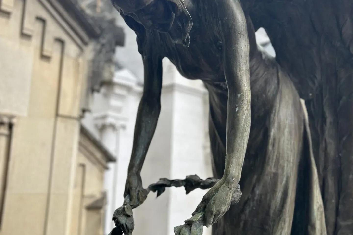 Primer plano de una escultura de bronce patinada de un ángel alado, inclinándose y colocando una corona de hojas sobre una...
