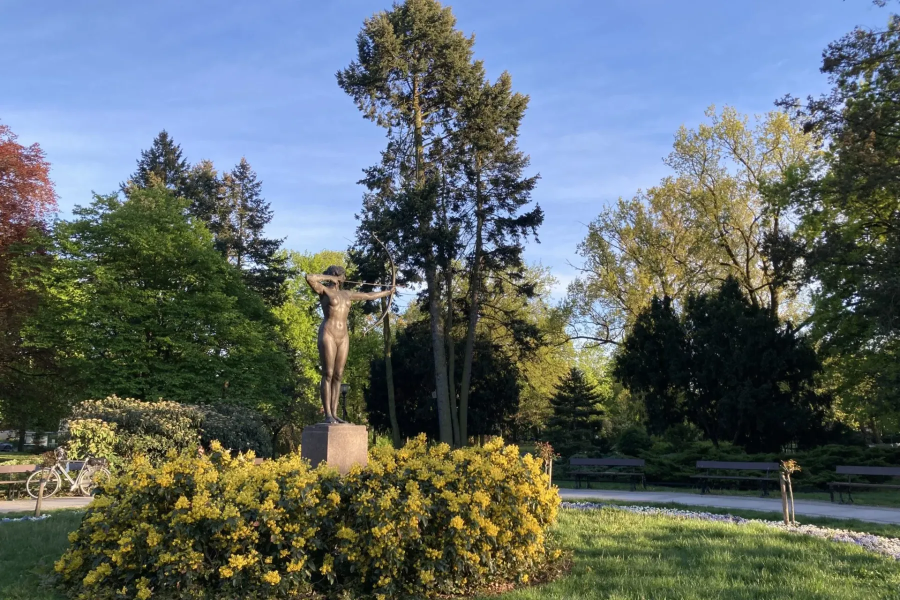 Bronze-Statue einer Bogenschützin in einem Park, umgeben von gelb blühenden Büschen, grünen Bäumen und Gras unter blauem H...