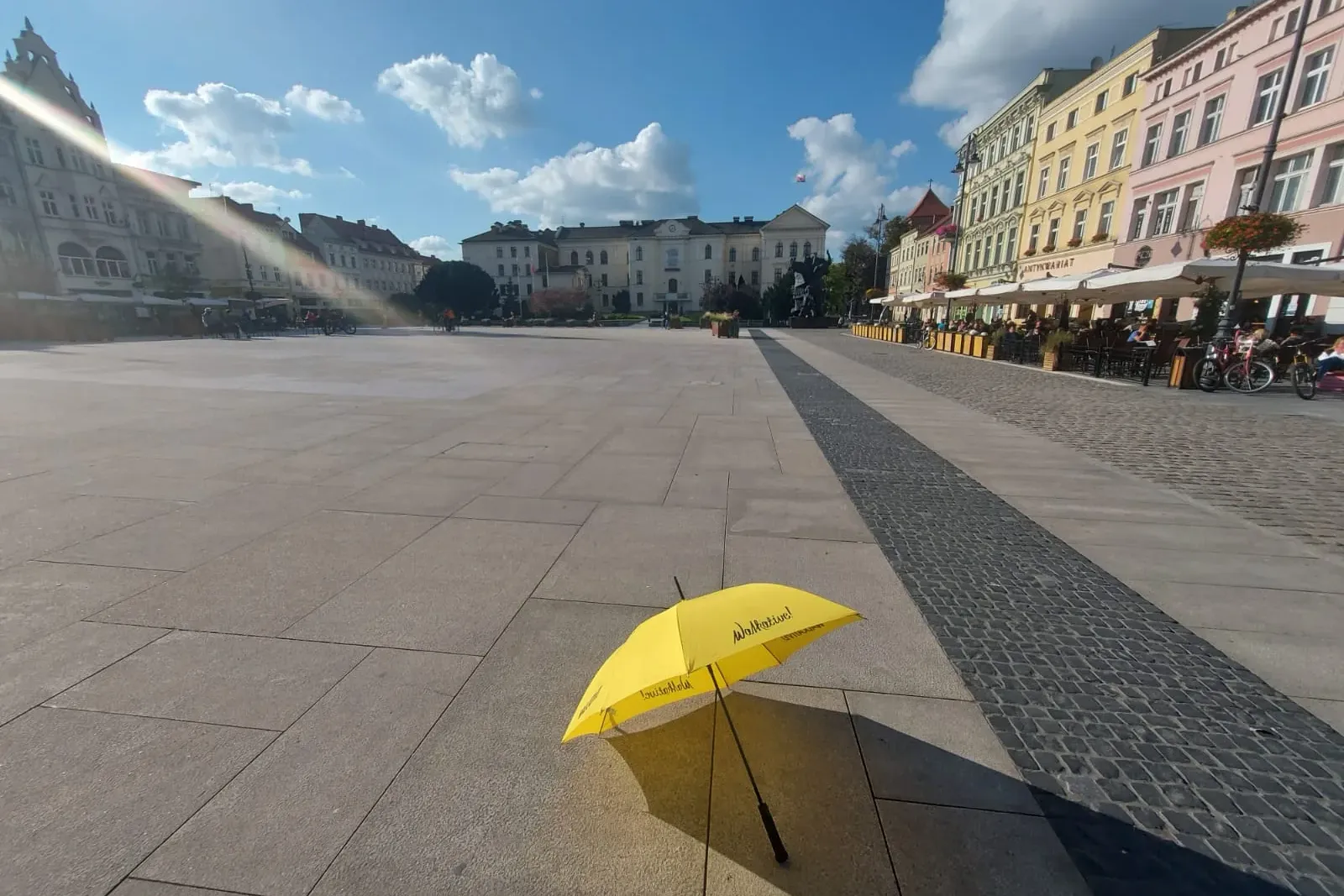 Żółta parasolka Walkative leży na dużym, pustym brukowanym rynku otoczonym zabytkowymi budynkami pod błękitnym niebem.