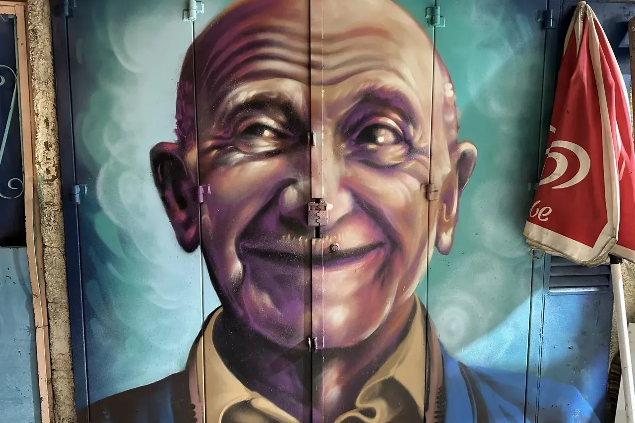 Mural colorido de un anciano calvo sonriente en puertas de metal azules, con un paraguas rojo plegado a la derecha.