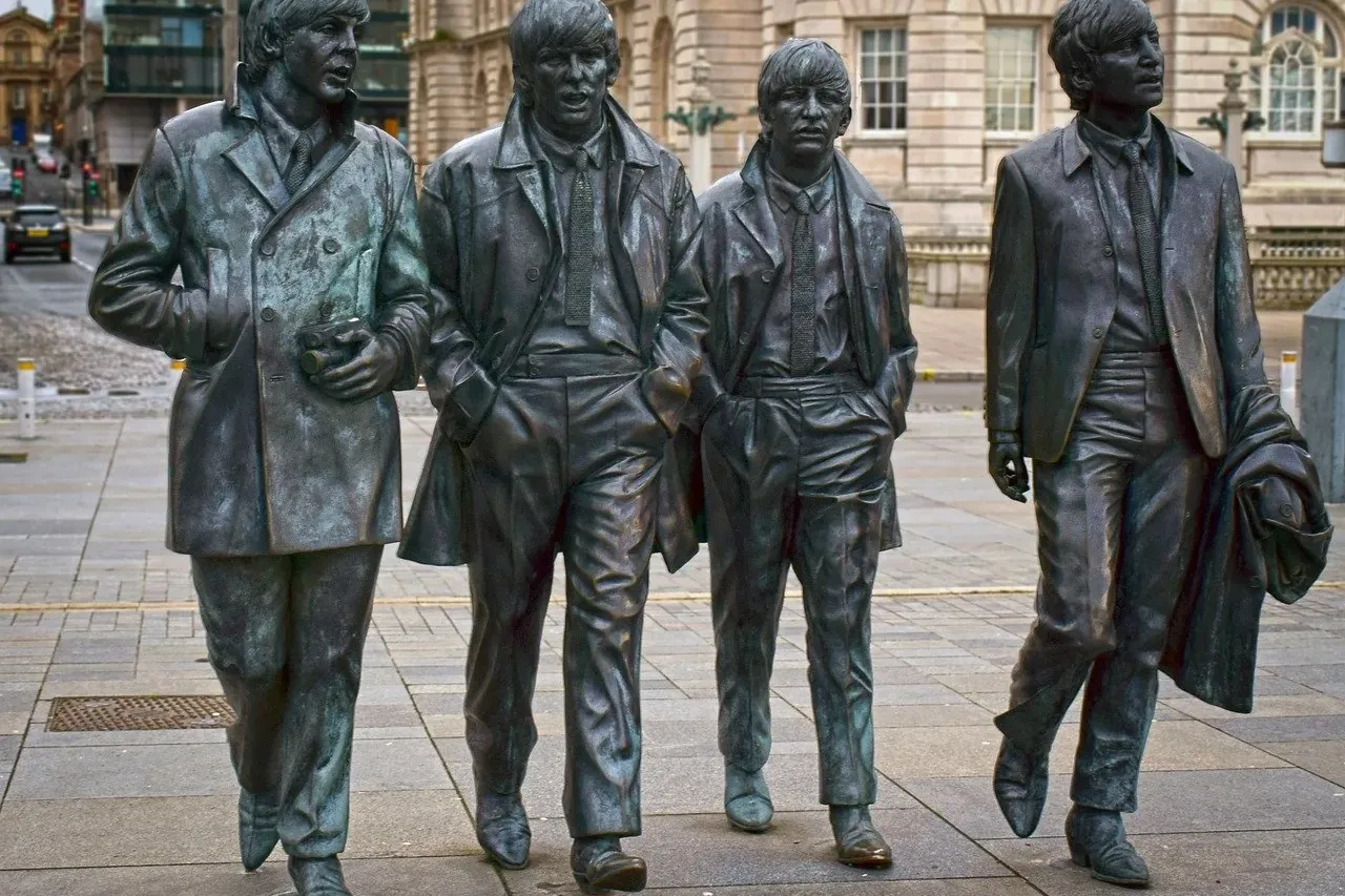 Bronze statues of The Beatles walking on a paved square in Liverpool, with historic stone buildings in the background.