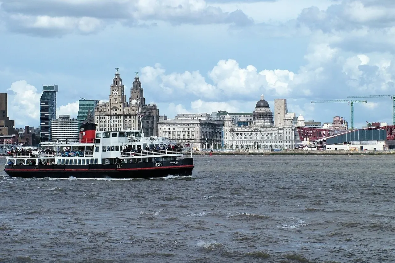 A ferry boat carrying passengers on rough water, with the Liverpool skyline and the Royal Liver Building visible under a c...