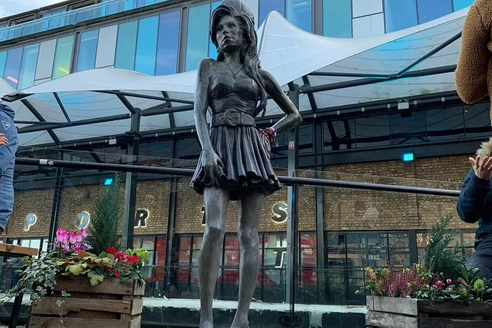 Brązowy posąg Amy Winehouse z ręką na biodrze, stojący na brukowanym placu przed nowoczesnymi budynkami pod zachmurzonym n...
