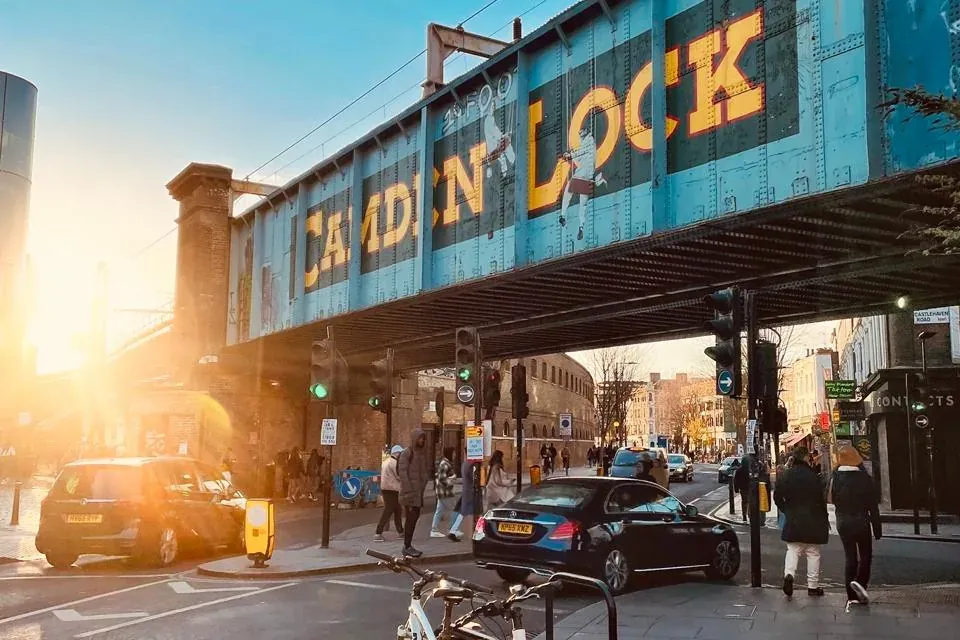 Niebieski most kolejowy z napisem "CAMDEN LOCK", przecinający ulicę z samochodami, pieszymi i zaparkowanymi rowerami o zac...
