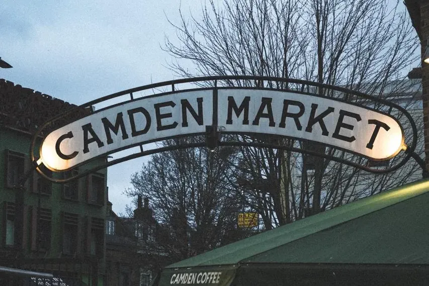 Oświetlony łukowy znak Camden Market o zmierzchu, z nagimi drzewami, budynkami i ludźmi na pierwszym planie.