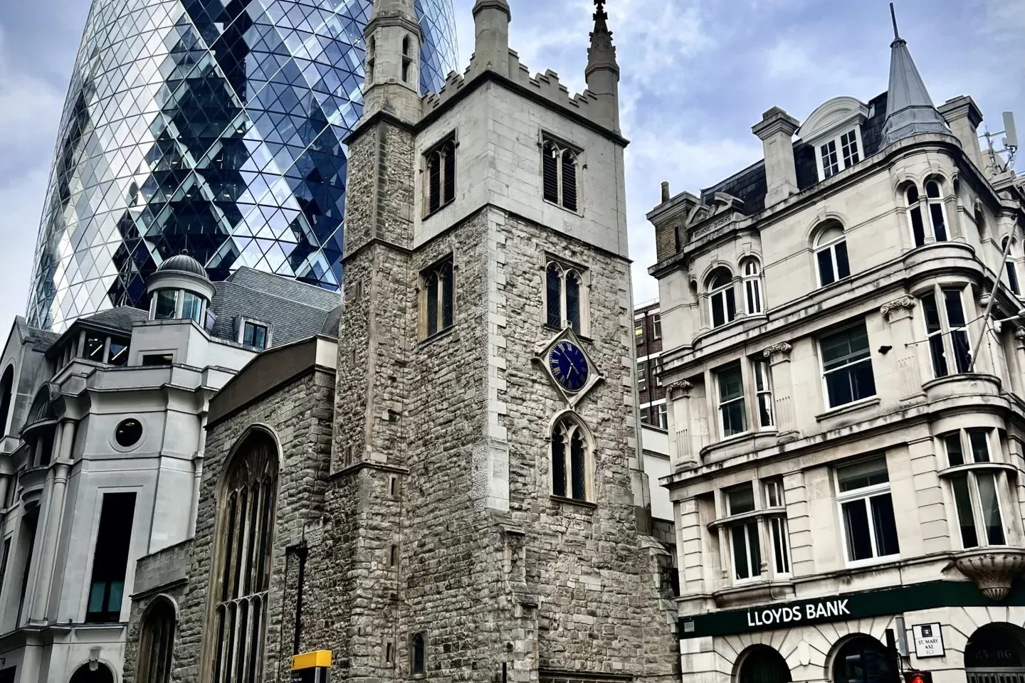 Der Gherkin-Wolkenkratzer, ein historischer Steinkirchturm mit blauer Uhr und ein Lloyds Bank Gebäude an einer belebten Lo...