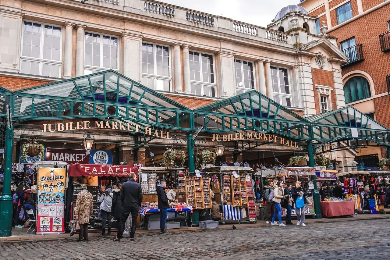 Hala targowa Jubilee Market Hall w Londynie z zielonym szklanym zadaszeniem nad straganami i ludźmi na brukowanej ulicy.