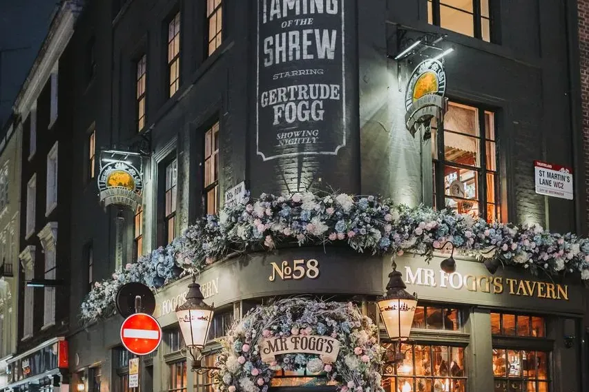 Zewnętrze Mr Fogg's Tavern w Londynie nocą, ozdobione niebiesko-różowymi girlandami kwiatowymi wokół wejścia i okien. Prze...