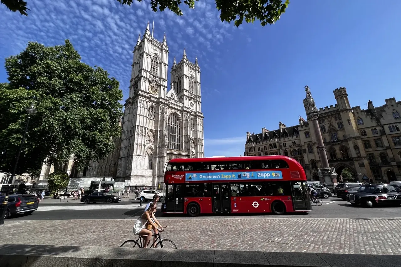 Westminster Abbey's historic facade and tower under a blue sky, with a red double-decker bus, cars, and people on the street.
