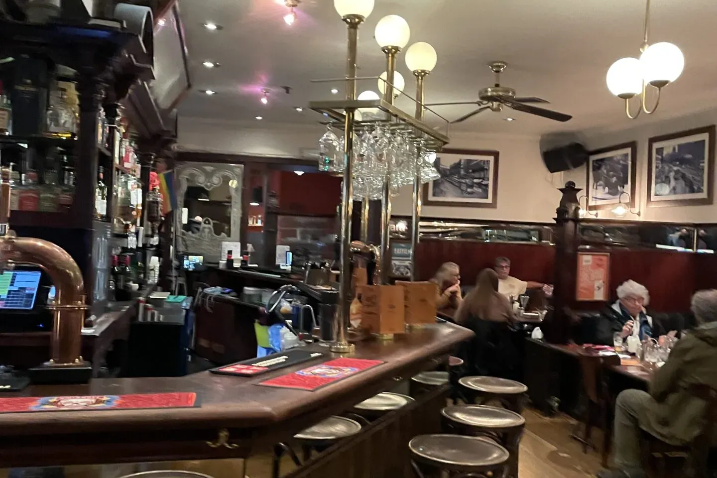 Interior de un pub tradicional con barra de madera, varios taburetes vacíos y personas sentadas en mesas.