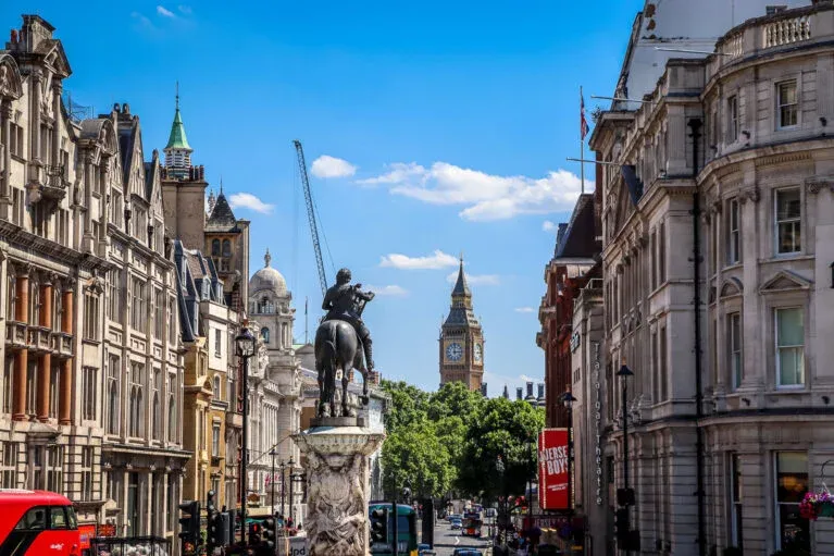 Equestrian statue in Trafalgar Square, London, with Big Ben visible in the distance down a busy street lined with buildings.