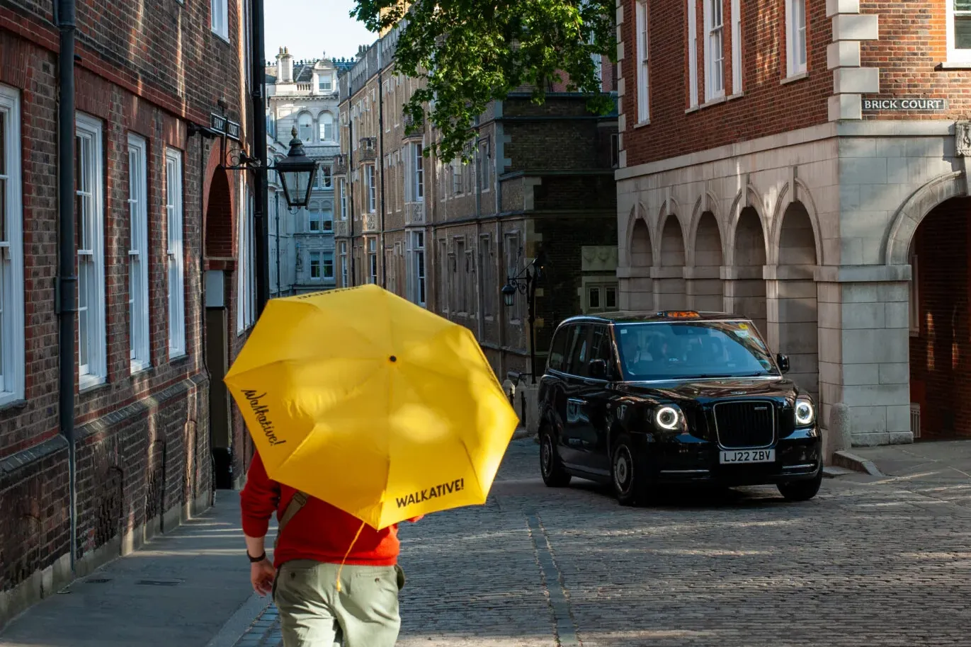 Person mit gelbem Regenschirm auf Kopfsteinpflasterstraße in Londons Brick Court, mit Backsteingebäuden und schwarzem Taxi.