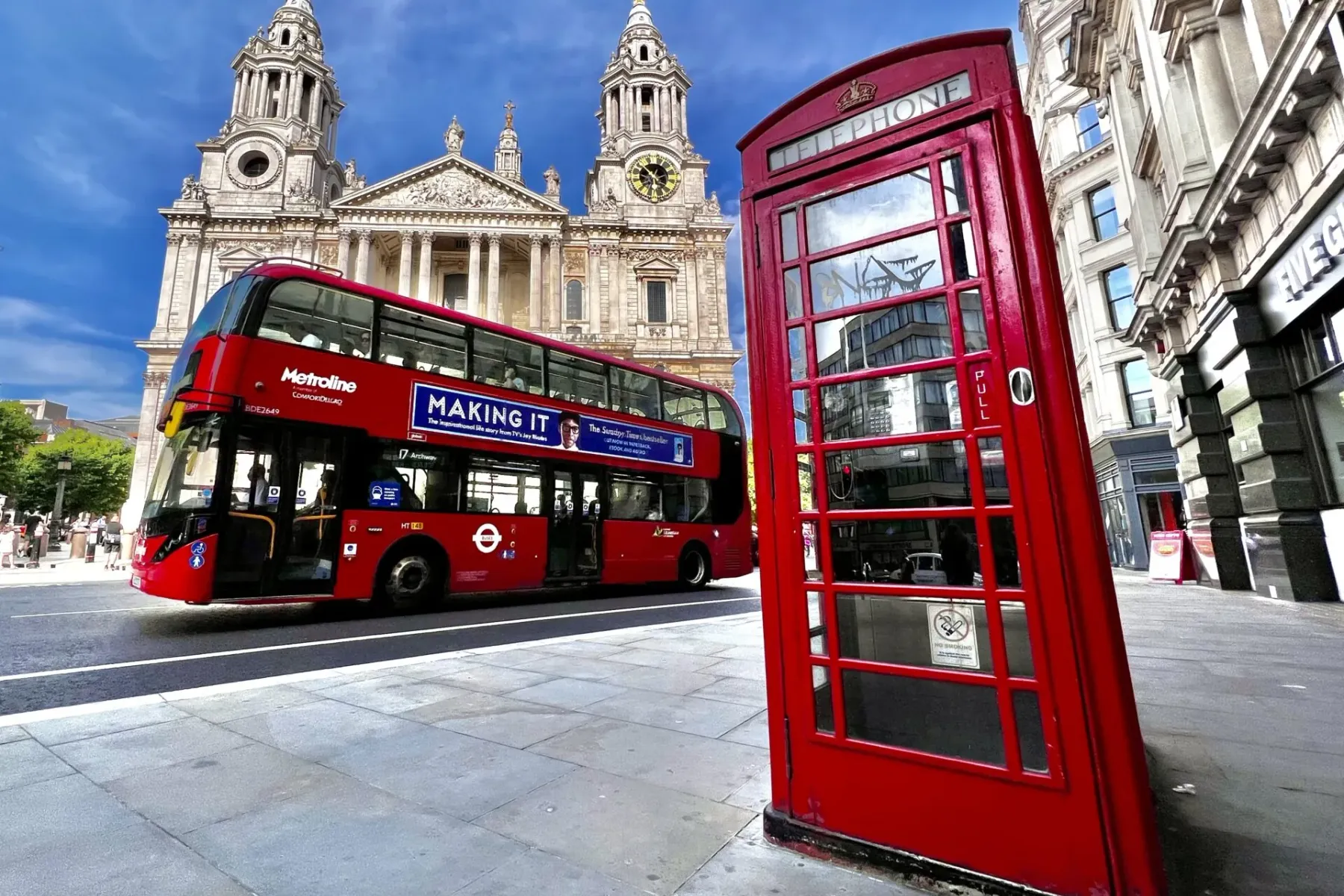 St. Paul's Kathedrale hinter einem roten Doppeldeckerbus und einer roten Telefonzelle auf einer sonnigen Londoner Straße.