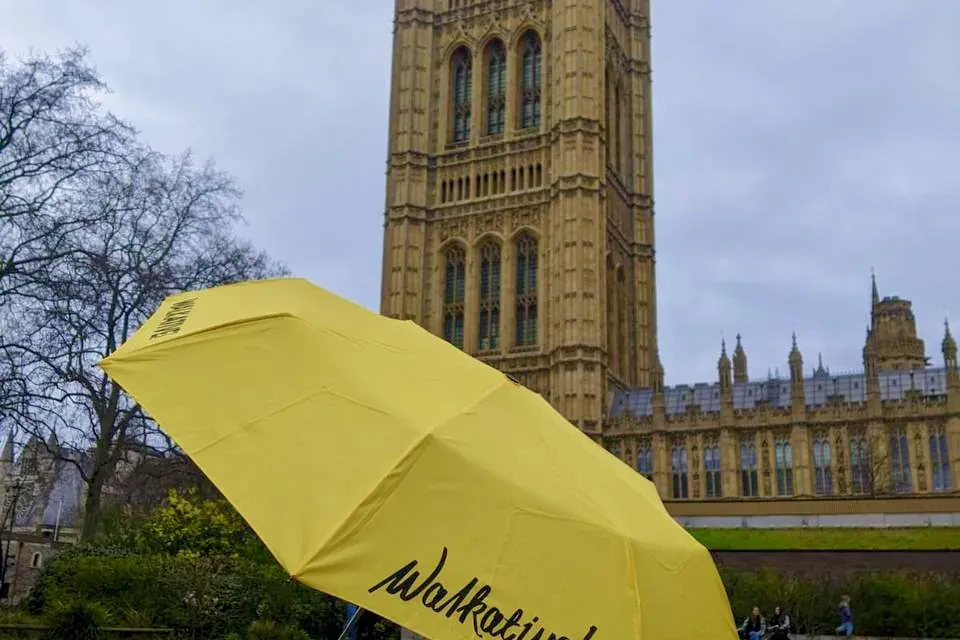 Yellow Walkative umbrella on a paved area with the Houses of Parliament and Victoria Tower in London visible behind it und...