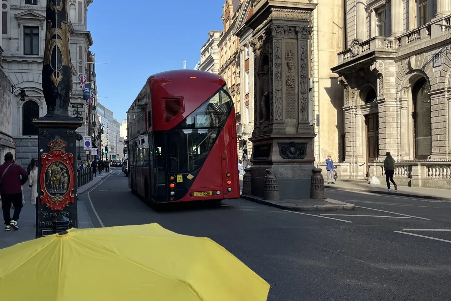 Gelber Walkative-Regenschirm im Vordergrund mit rotem Doppeldeckerbus, Drachenstatue und historischen Gebäuden auf einer S...