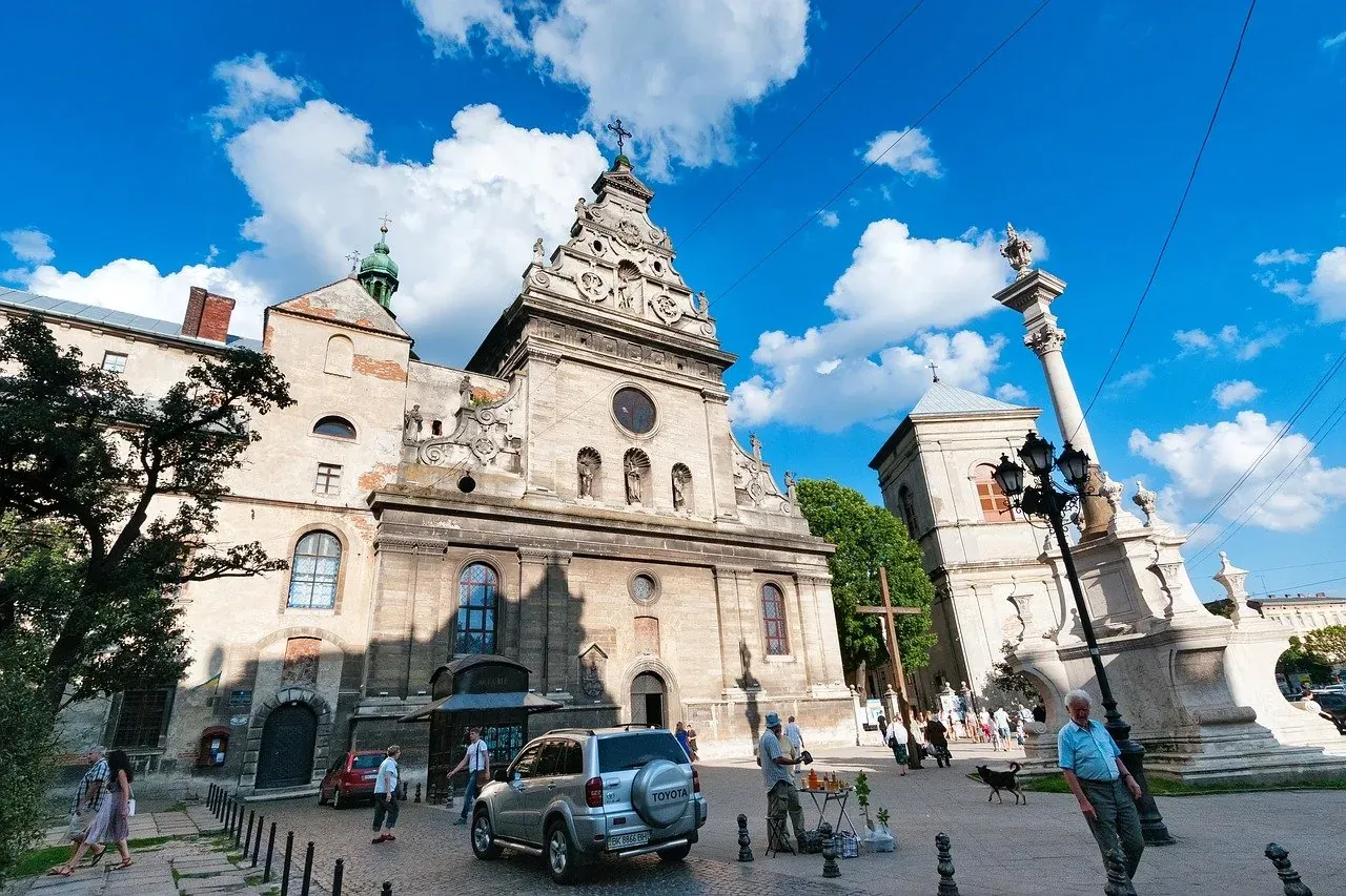 Iglesia barroca de San Andrés en Lviv con una alta y ornamentada Columna de Juan de Dukla en un día soleado. Gente caminando.