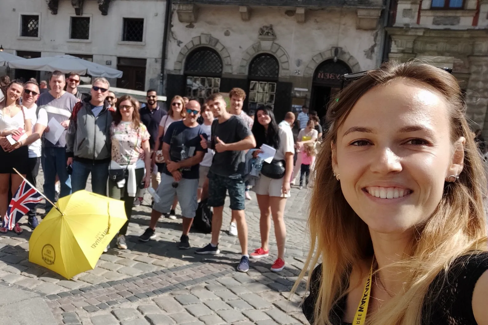 Mujer sonriente se toma un selfie con un grupo de turistas en una calle adoquinada de Lviv. Un paraguas amarillo y la band...