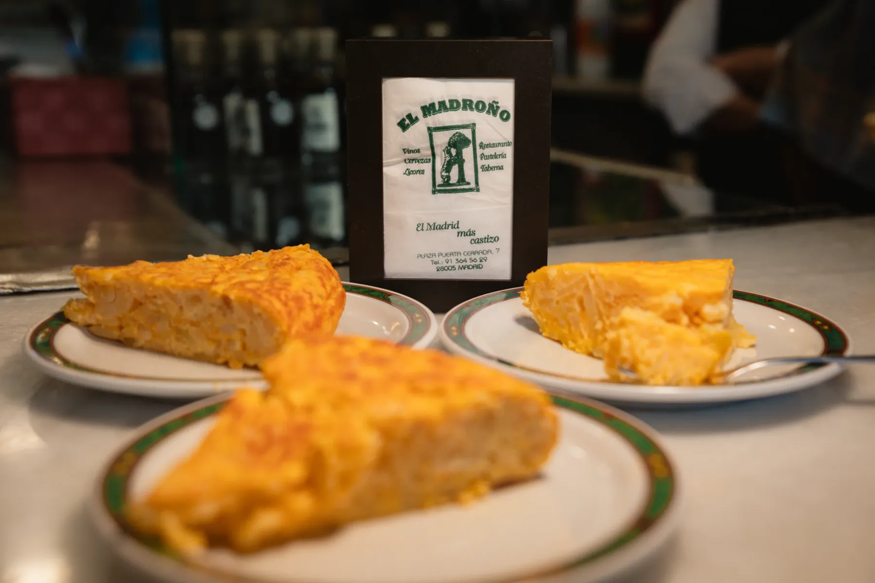 Three slices of Spanish tortilla on white plates with green and brown rims, with a framed napkin from 'El Madroño' in the ...
