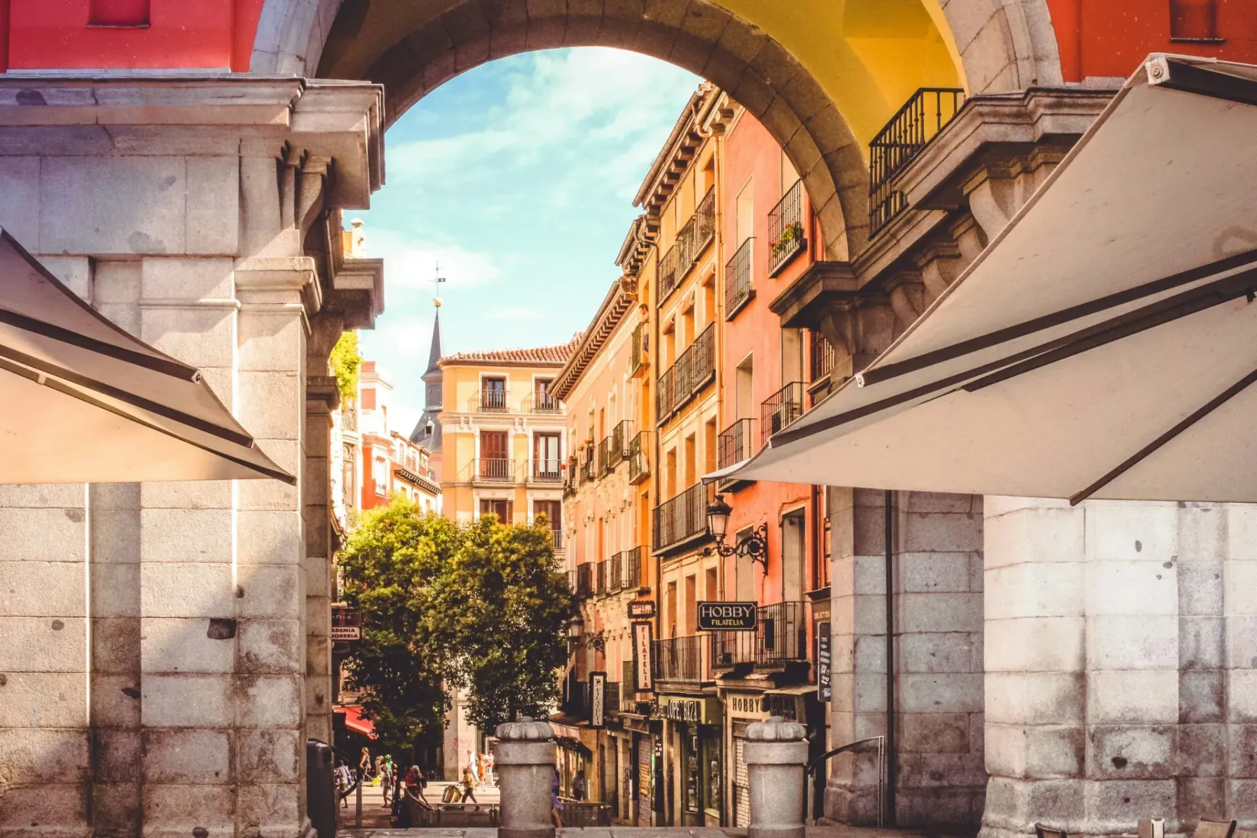 View through a large stone archway in Madrid, showing a street with colorful buildings, balconies, and a distant spire, un...