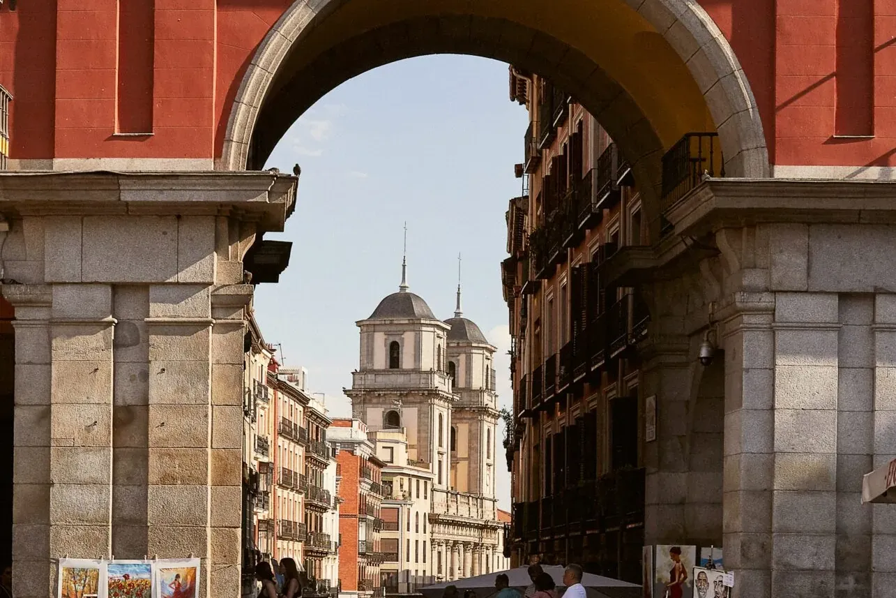 Street artists display paintings near a stone archway in Madrid's Plaza Mayor, with church towers visible in the distance.
