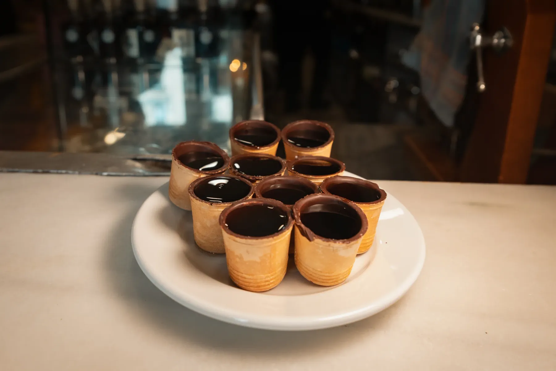 Nine small edible cups with chocolate-dipped rims filled with dark liquid, arranged on a white plate.
