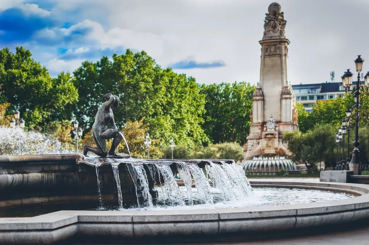 Una escultura de bronce arrodillada sobre una fuente escalonada, con agua fluyendo. Al fondo, un alto monumento de piedra ...