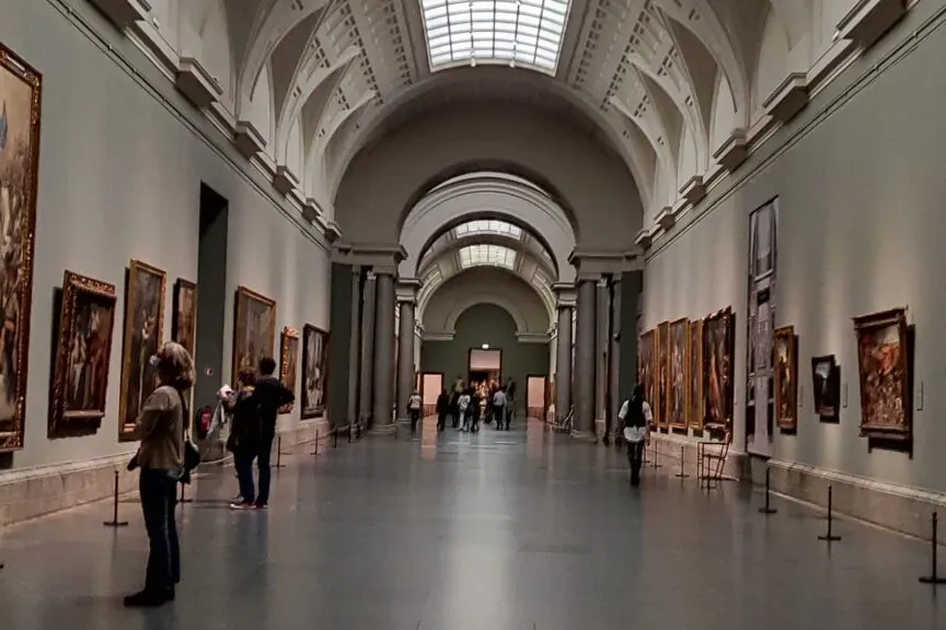 Largo y alto pasillo de museo con cuadros enmarcados en ambas paredes y una gran claraboya rectangular en el techo. Person...