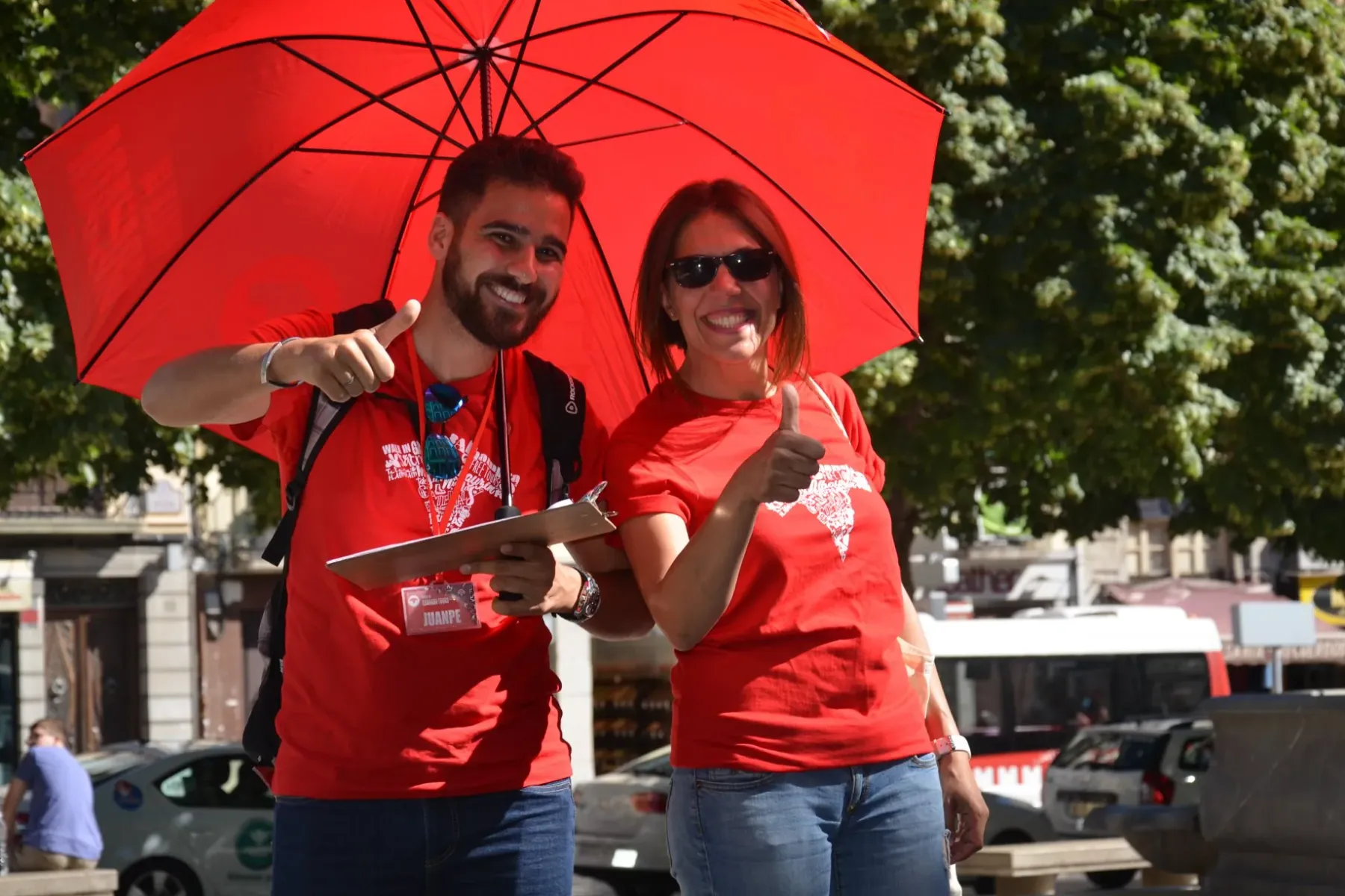 Two smiling Walkative tour guides, a man and a woman, stand under a red umbrella, giving thumbs-up on a sunny street.