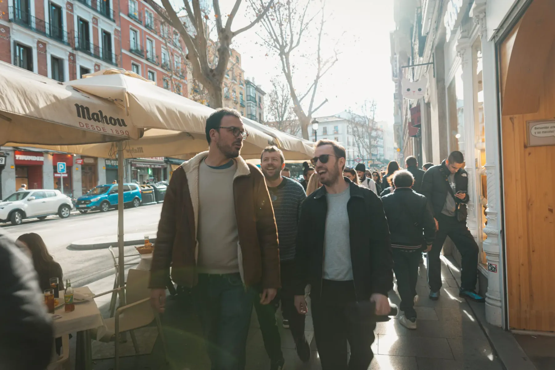Two men smiling and walking on a sunny Madrid street with other pedestrians, buildings, and a cafe awning visible.