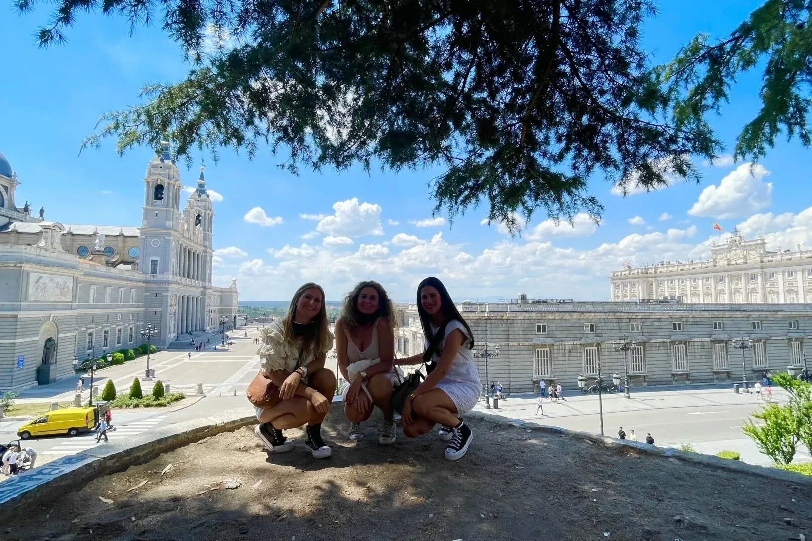 Tres mujeres en cuclillas frente a la Catedral de la Almudena y el Palacio Real de Madrid bajo cielo azul.