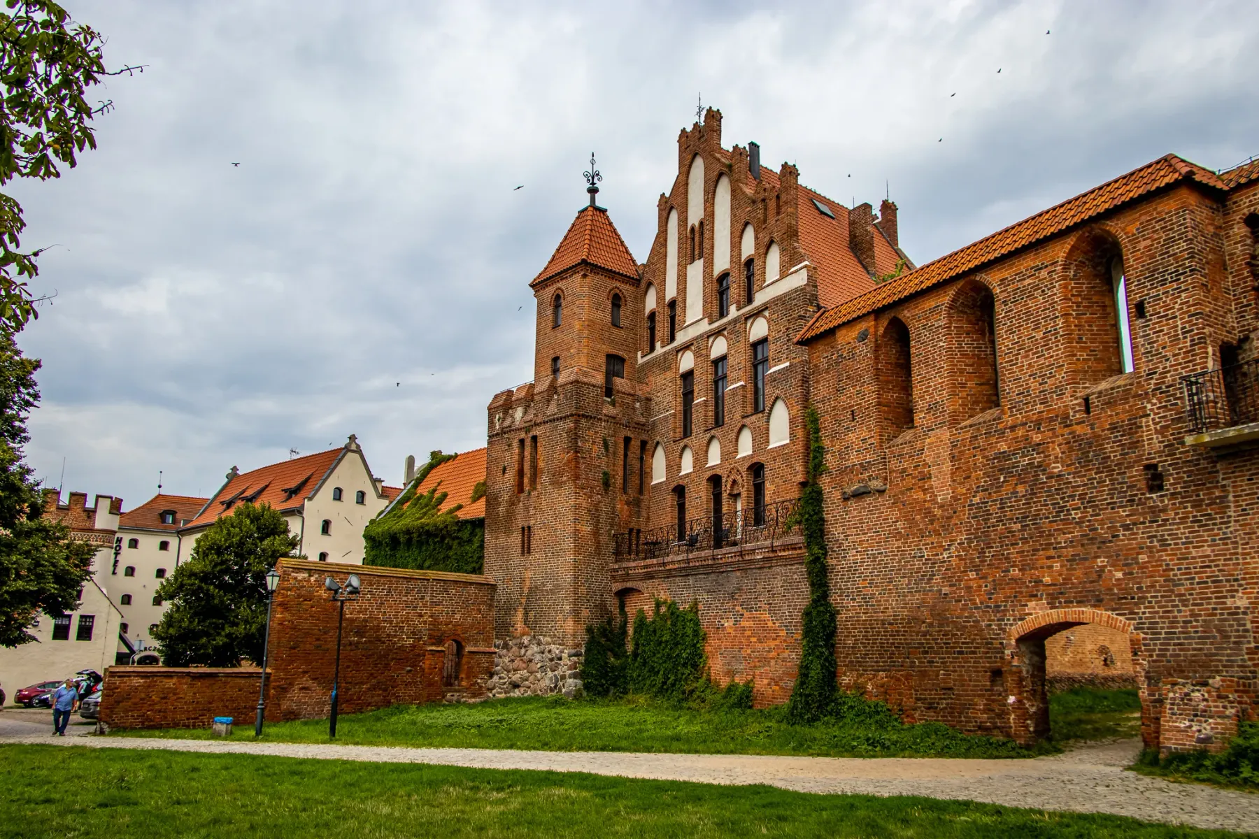 Gotische Backsteinburg mit markantem Turm, teilweise Ruinen mit Bogenöffnungen, grünem Rasen und bewölktem Himmel.