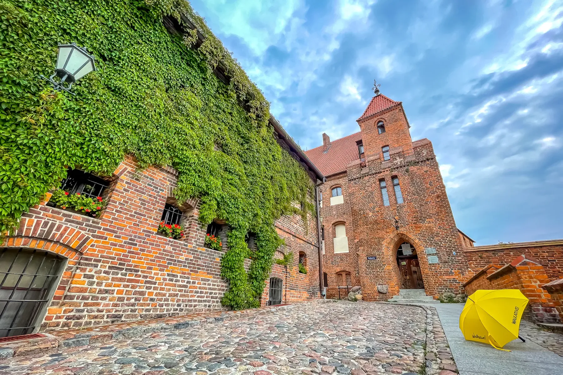Kopfsteinpflasterstraße führt zu einem historischen Backsteingebäude, das teilweise mit grünem Efeu bedeckt ist, unter bew...
