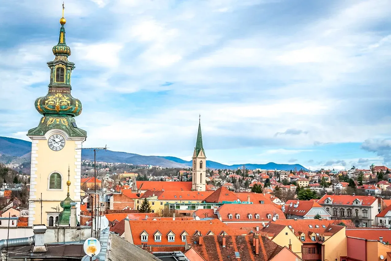 Vista del casco antiguo de Zagreb con una torre de reloj barroca, numerosos tejados rojos y colinas lejanas.