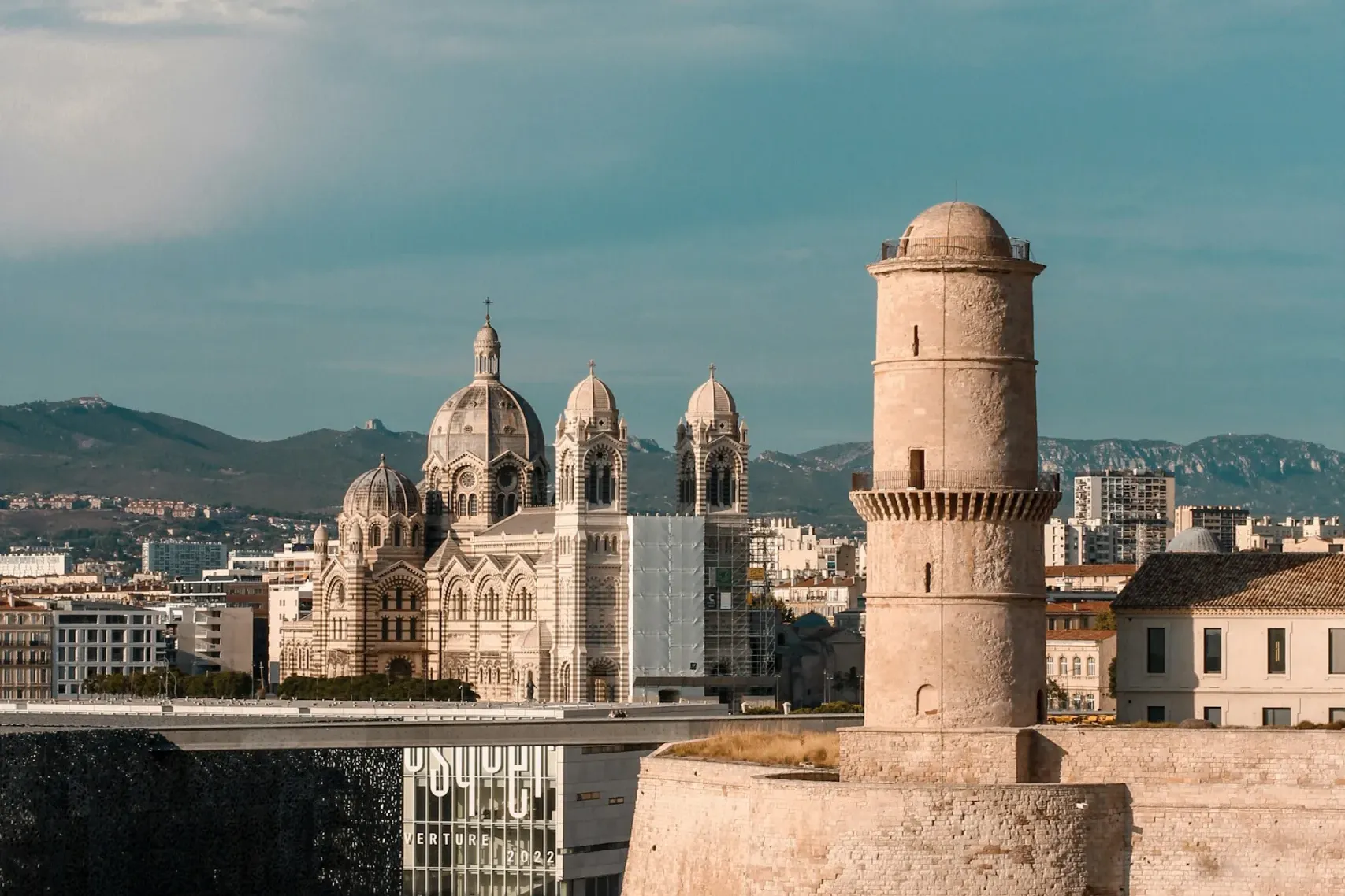 Torre y muros del Fuerte Saint-Jean de Marsella junto al mar, con la Catedral La Major, museo Mucem y montañas lejanas.