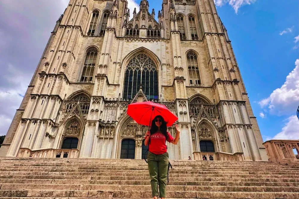 Una mujer con paraguas rojo de pie en anchas escaleras de piedra frente a la gran Catedral gótica de San Miguel y Santa Gú...