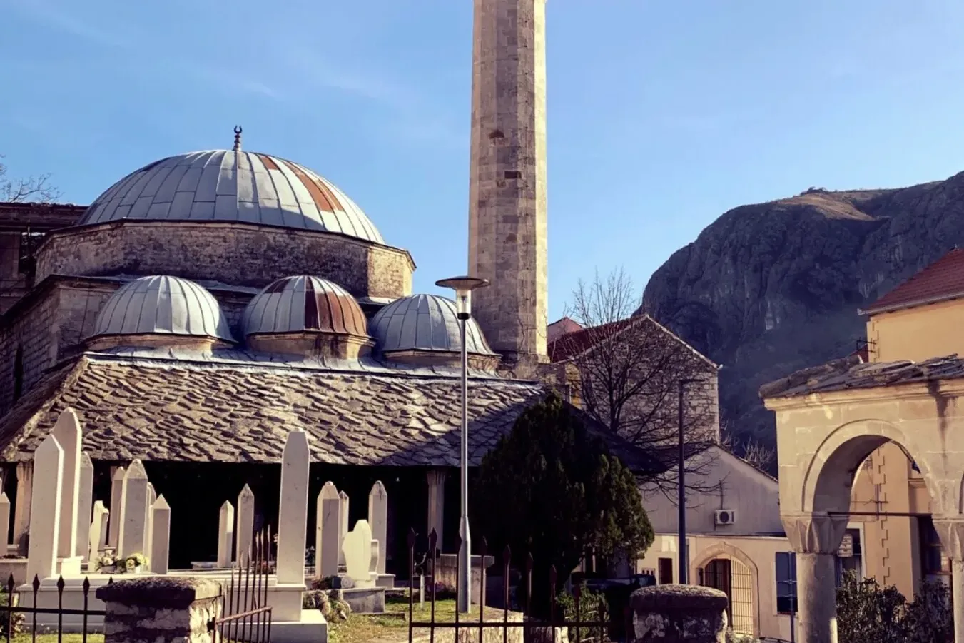 Kamienny meczet z wieloma kopułami i wysokim minaretem, otoczony cmentarzem z białymi nagrobkami pod błękitnym niebem.